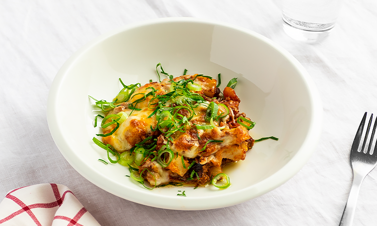 Enkel moussaka med grønnsaker og vegetardeig servert i en dyp hvit skål med en gaffel på høyre side og et glass vann i bakgrunnen