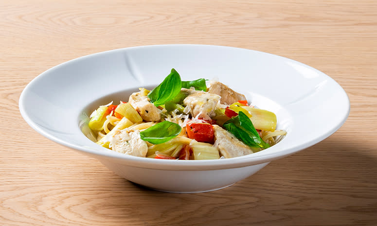 Pasta med kylling, paprika og purre toppet med basilikum, servert i en hvit skål med brede kanter på en tre overflate