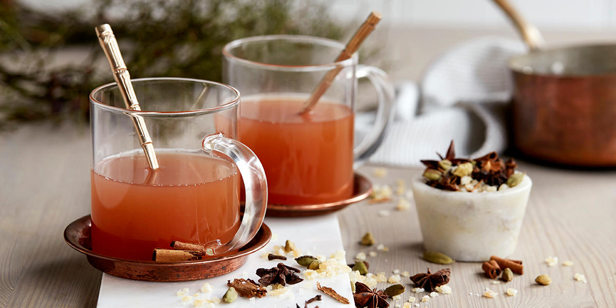 Eplegløgg med hjemmelaget krydderblanding servert i gjennomsiktige glasskopper, på messingskåler. Koppene står på lyst trebord med granbar i bakgrunnen, og en liten marmorskål med krydderblanding ved siden av. Litt av krydderblandingen er strødd rundt.