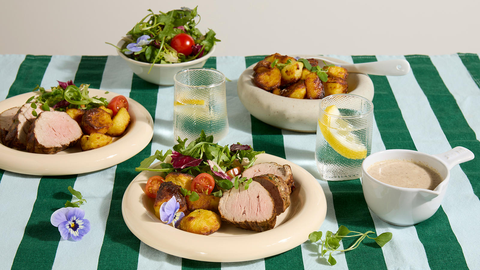 Indrefilet av svin med crispy ovnsbakte poteter og soppsaus servert på lyse tallerkner på grønnstripete duk. Skål med crispy ovnsbakte poteter og frisk salat i bakgrunnen.