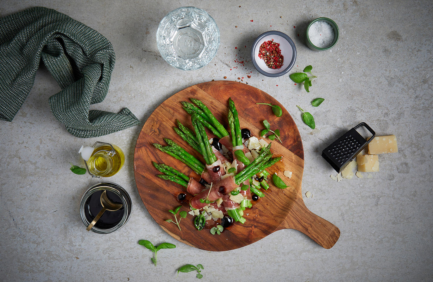 Asparges med spekeskinke og parmesan servert på rund trefjøl med revet parmesan, balsamico og frisk basilikum. Små skåler med tilbehør rundt.