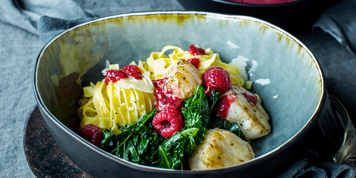 Pasta med kamskjell, smørdampet grønnkål, bringebærvinaigrette og ferske bringebær, servert i en blålig rustikk skål, på mørk linduk. Revet parmesan er drysset over.