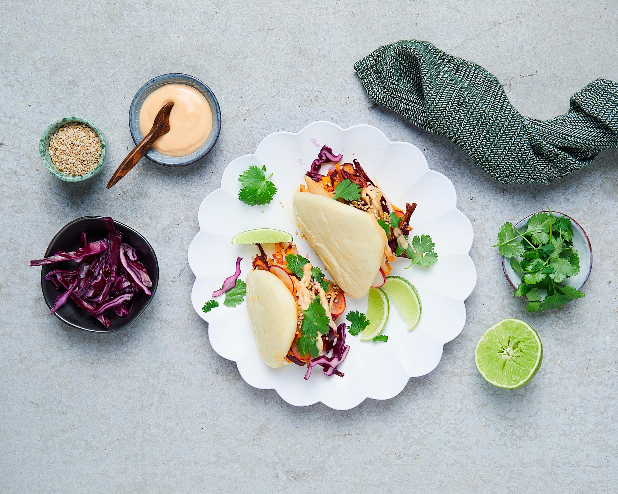 2 stk koreanske kyllingklubber med coleslaw på en hvit tallerken. Rundt står ulike skåler med sesamfrø, chili mayo, rødkål, koriander og lime. 