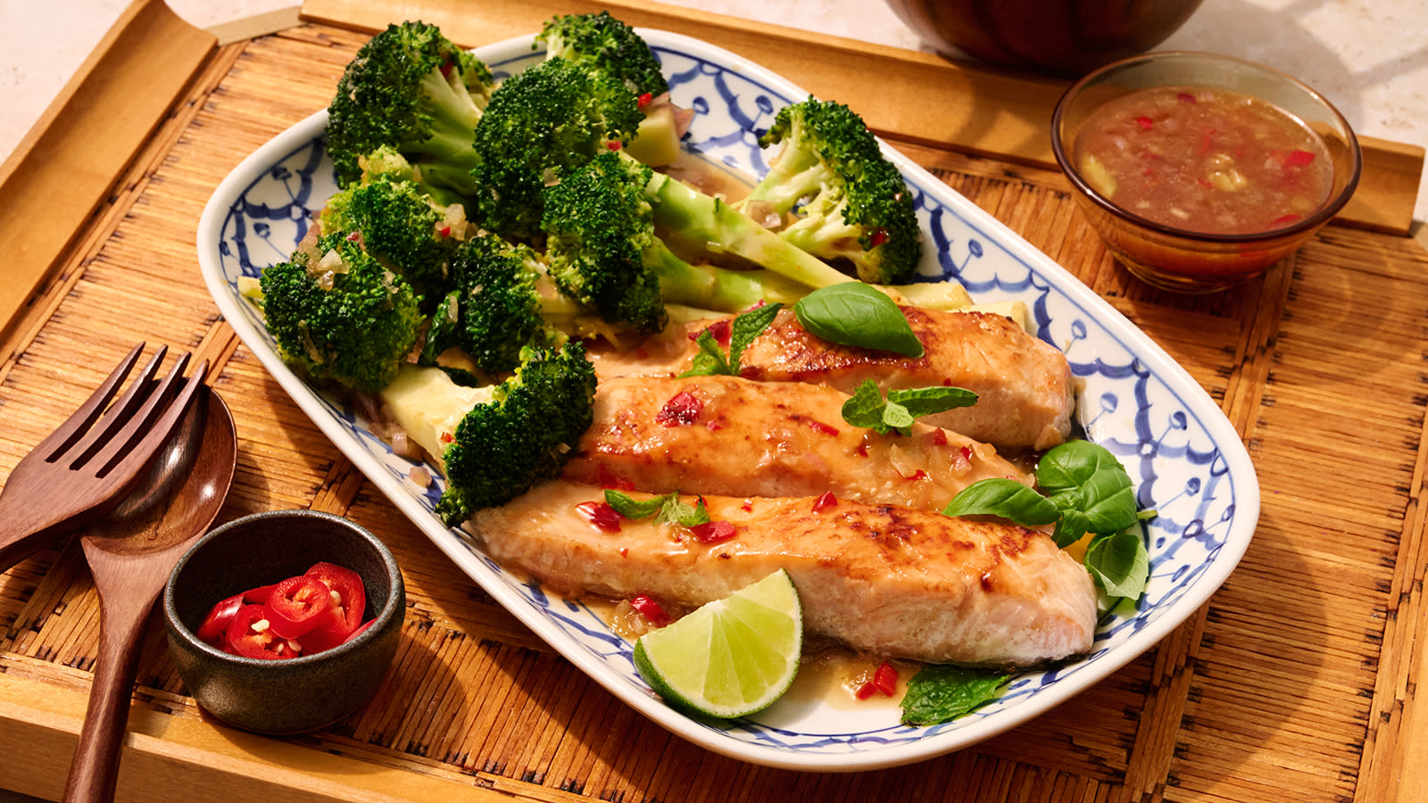 Laks Sam Lott med brokkoli servert med syrlig chilisaus, limebåt og friske urter på avlangt hvitt- og blåmønstret fat.