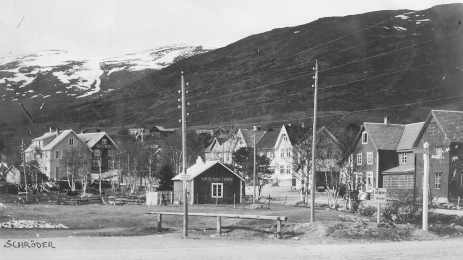 Svart hvitt bilde av gammel gate med hus i flere sjateringer. Snødekkede fjell i bakgrunnen. 