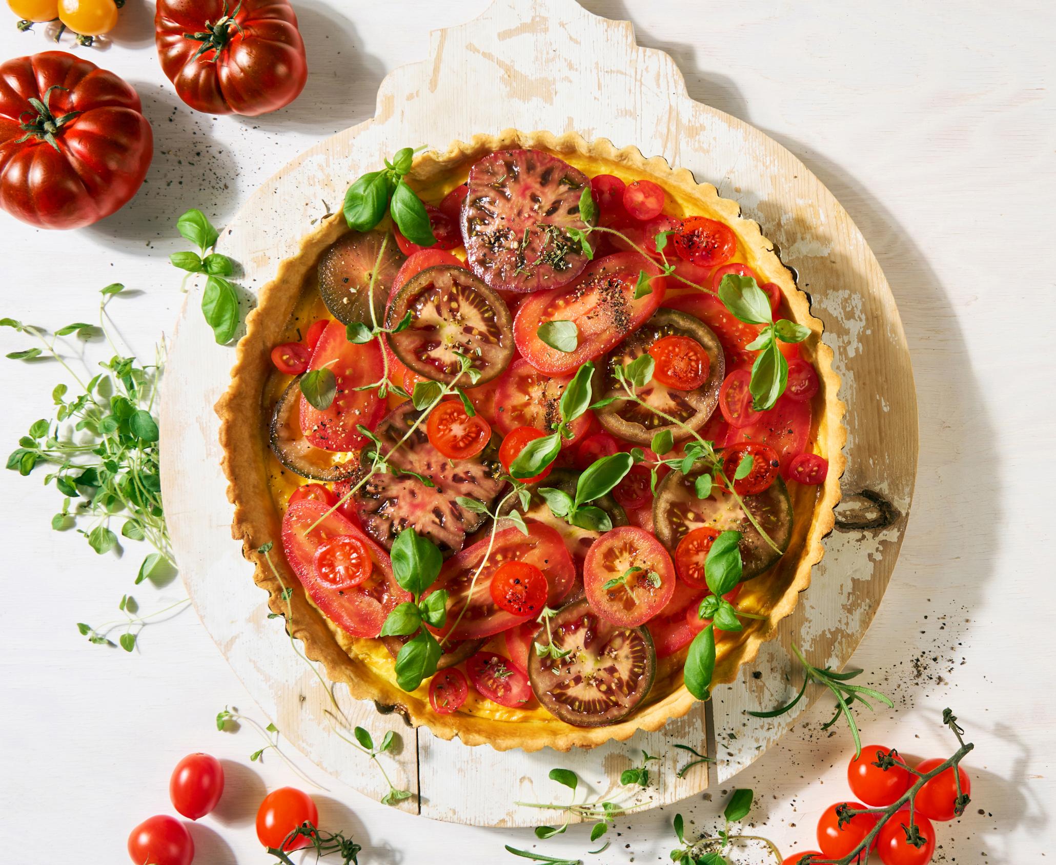 Frisk tomatpai med hjemmelaget paibunn og eggestand toppet med friske urter, og servert på rund rustikk trefjøl med hvit maling på hvit bakgrunn. Friske tomater i flere farger og fasonger, og friske urter rundt.