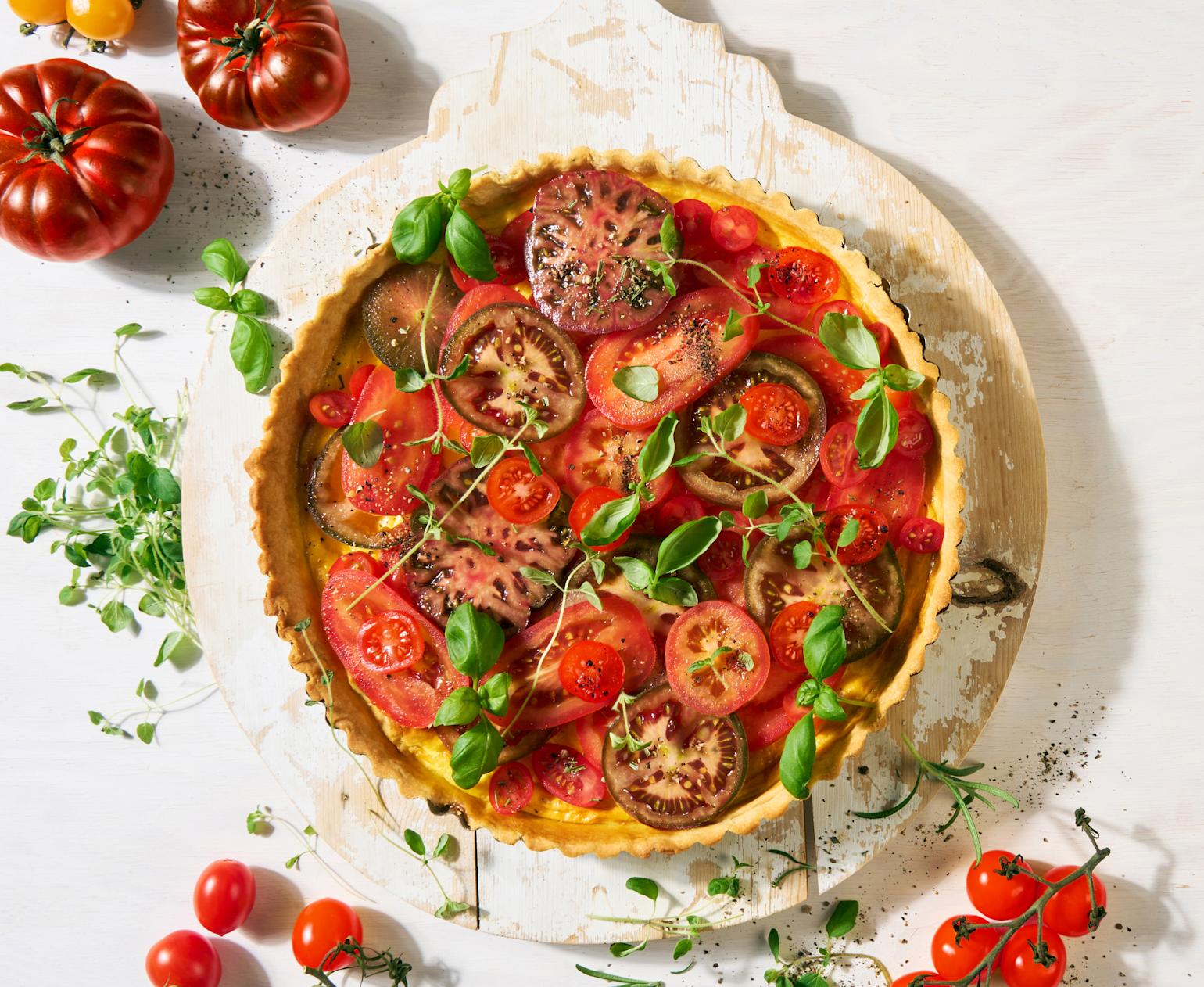 Frisk tomatpai med hjemmelaget paibunn og eggestand toppet med friske urter, og servert på rund rustikk trefjøl med hvit maling på hvit bakgrunn. Friske tomater i flere farger og fasonger, og friske urter rundt.
