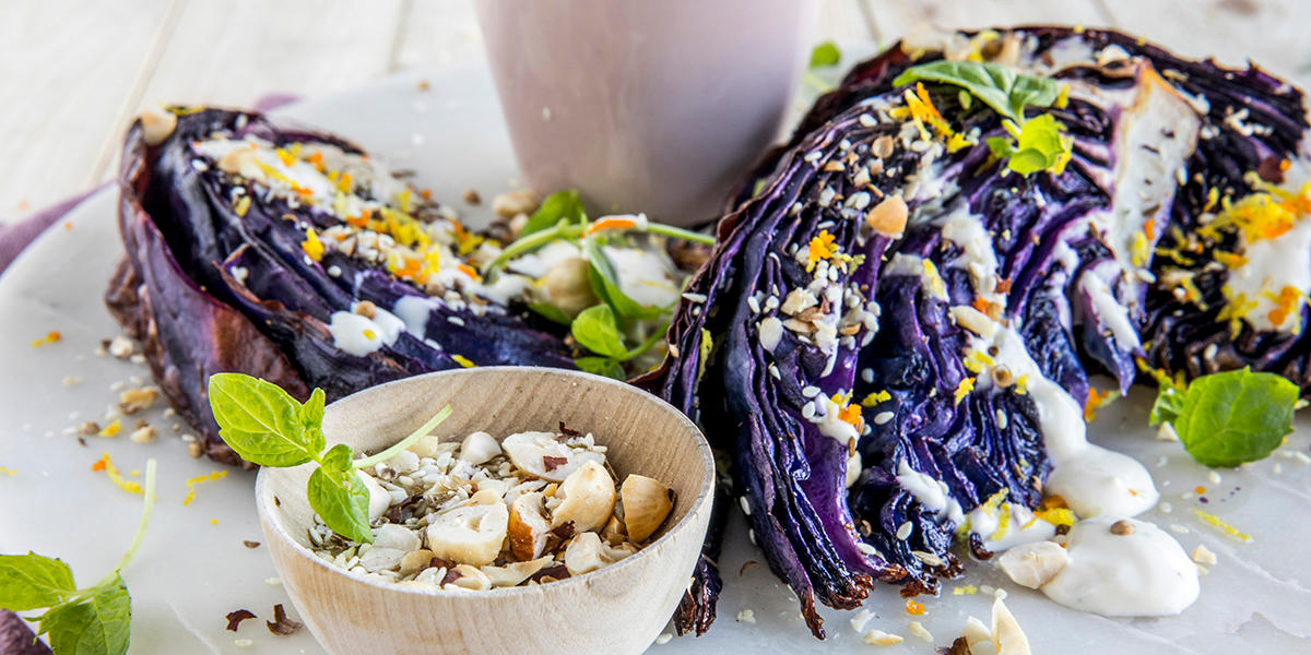 Pannestekt rødkål med dukkah og yoghurtdressing servert på hvitt marmorfat overstrødd med ferske urter. Liten treskål med dukkah på siden.