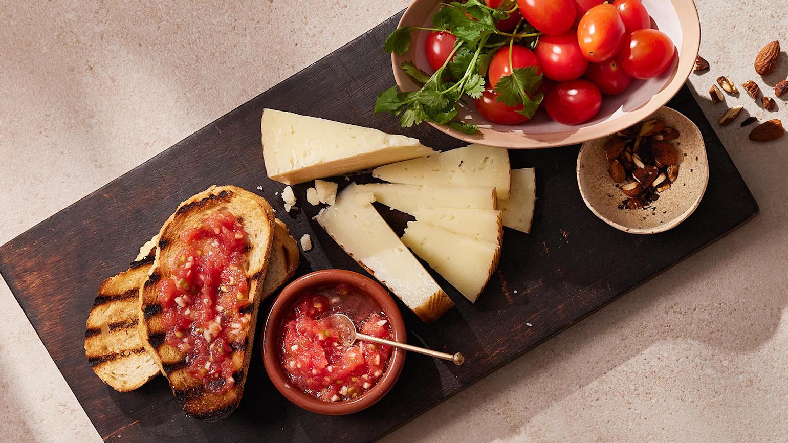 Pan Catalan - Spansk tomatbrød servert på m ørk trefjøl med ost og en skål tomater.