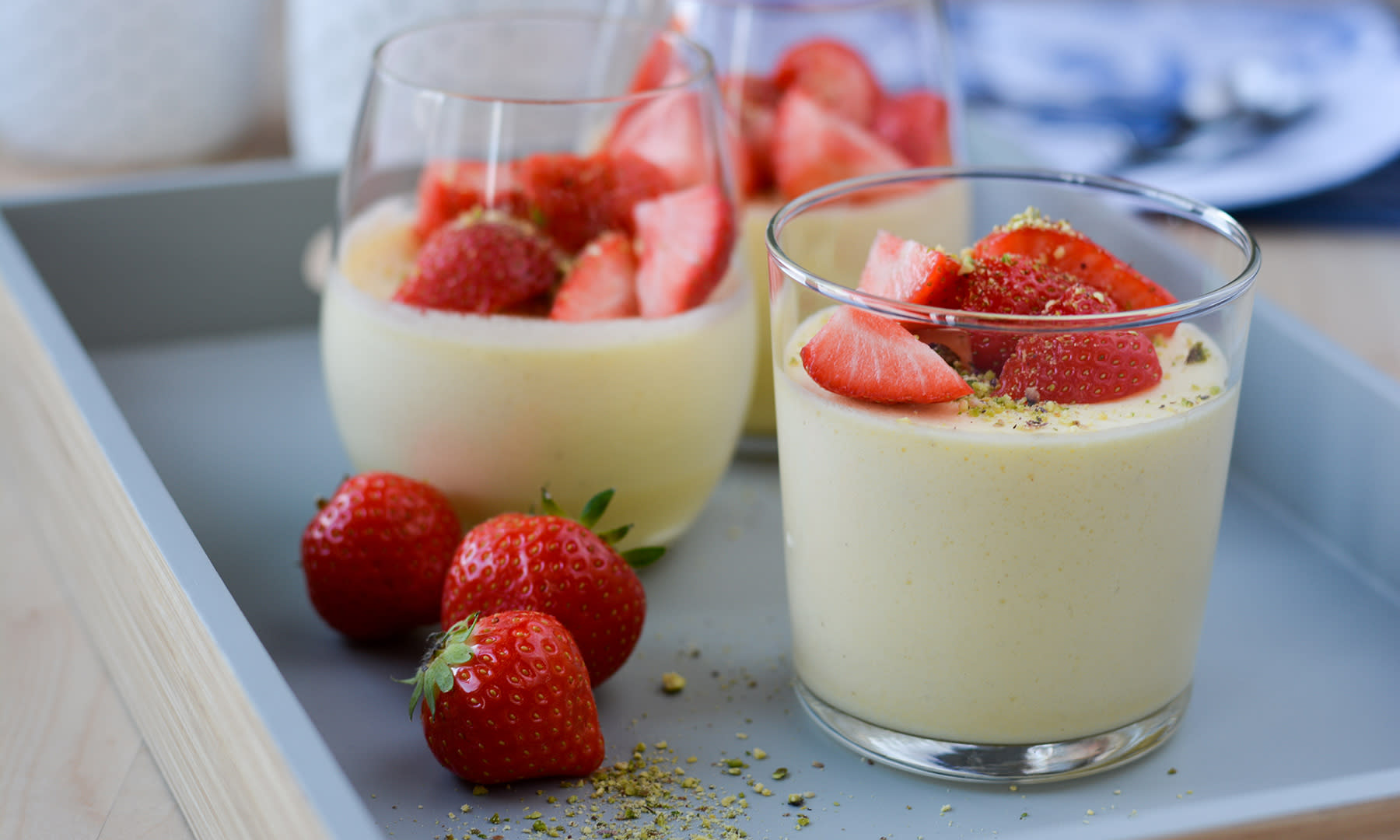 To glass med klassisk vaniljefromasj pyntet med jordbær og hakkede pistasjnøtter, servert på blåmalt serveringsbrett i tre. Ferske jordbær ved siden av.