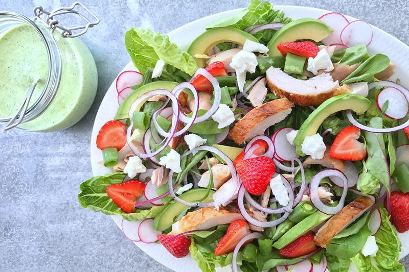 Lekker kyllingsalat med jordbær og urtedressing, rødløk, avokado, redikk og fetaost, servert på hvitt fat med krukke med urtedressing ved siden av.