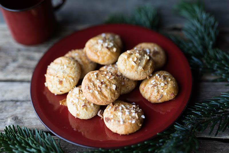 Serinakaker toppet med nøtter og perlesukker, servert på julerødt fat på rustikk trebord, omringet av granbar. Rød kopp i bakgrunnen.