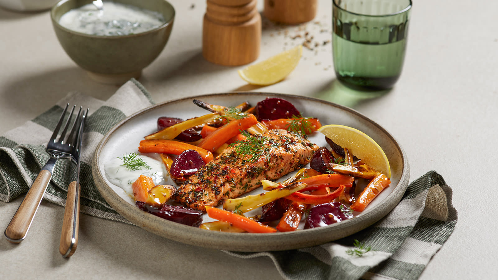 Ovnsbakt laks med ovnsbakte rotgrønnsaker og dillyoghurt servert på lys keramikkasjett på grønnstripete kjøkkenhåndkle. Skål med dillyoghurt, og grønt vannglass i bakgrunnen.