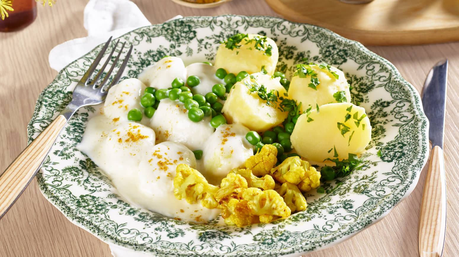 Fiskeboller med karristekt blomkål servert på grønnmønstret tallerken med hvit saus, erter og kokte poteter.