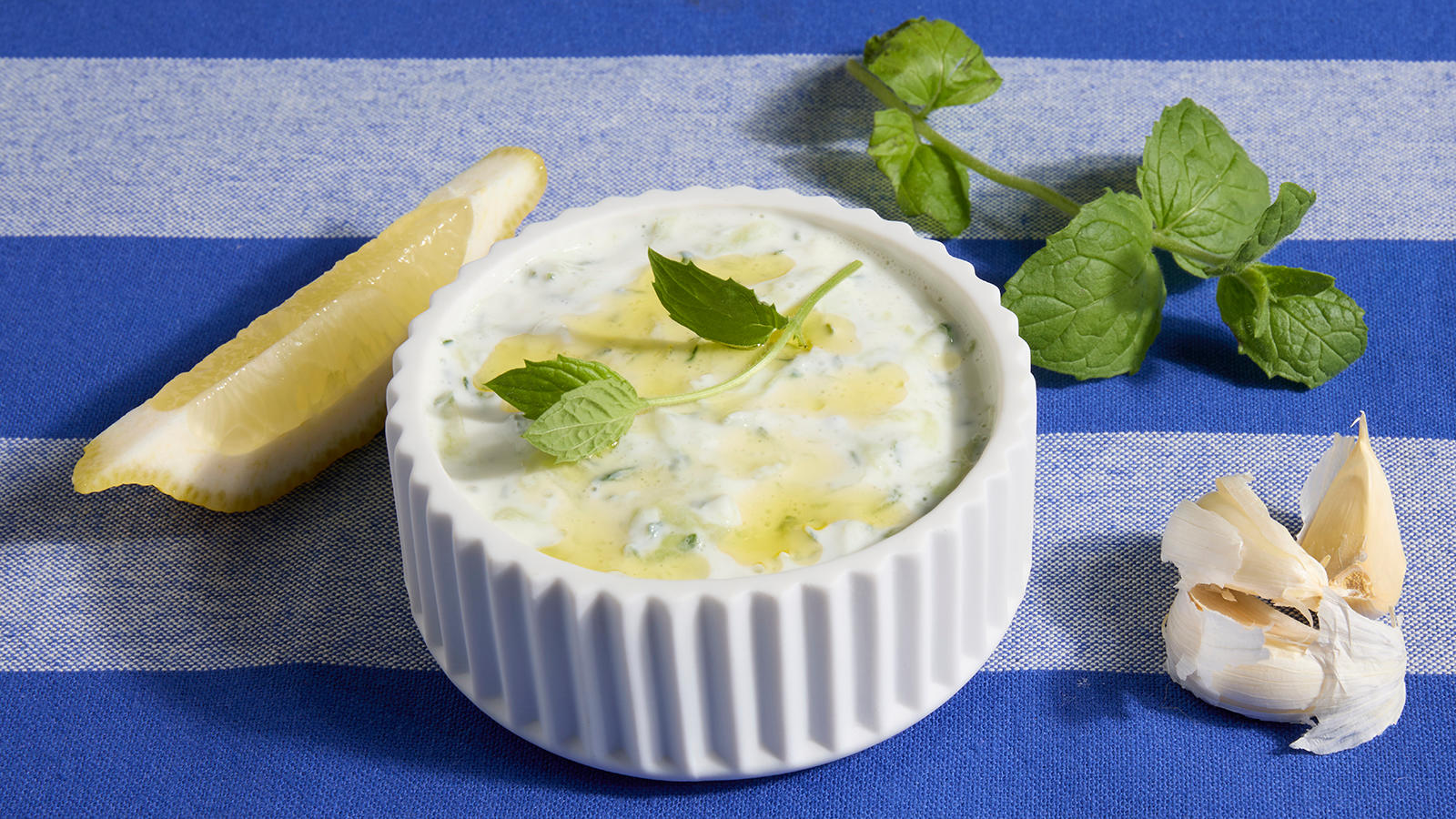 Hjemmelaget tzatziki servert i hvit skål med urter og olivenolje på toppen. Sitronbåt, urter og hvitløk ved siden av.