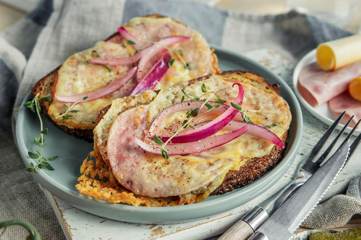 To ostesmørbrød med ost og skinke, servert på blågrønn tallerken og blåstripete linduk. Toppet med sitronsyltet rødløk og fersk timian.