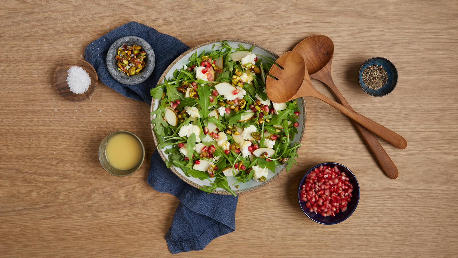 Høstsalat med pære, ruccola, chevre og pistasjkrønsj servert i grå bolle med små skåler dressing, havsalt, pistasjenøtter, granateplefrø og pepper på siden.