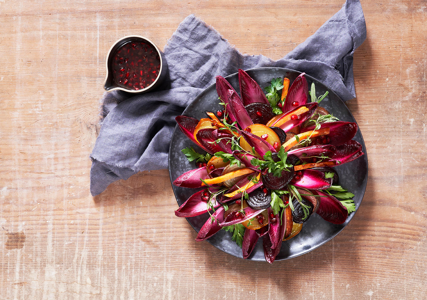 Bakt høstsalat med beter, rød raddiciosalat, bakte rotgrønnsaker og grønne urter. Ved siden av står en skål med dressing med granateplekjerner.