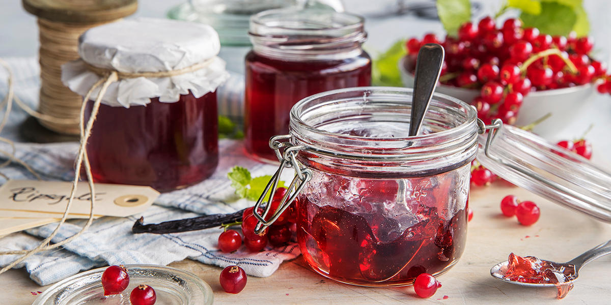 Tre glass med ripsgelé servert på rustikk trebord med ferske rips omkring, og i en skål i bakgrunnen. Hyssing, navnelapper og syltetøykleder i bakgrunnen.