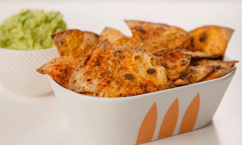 Ovnsbakte speltlompechips med krydder servert i hvit serveringsbolle med oransje mønster. Bolle med guacamole i bakgrunnen.