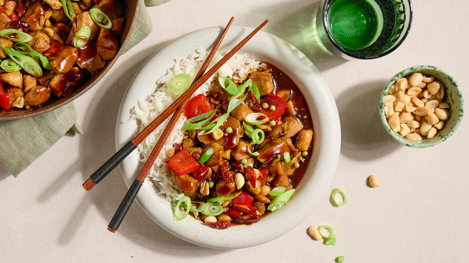 Kung Pao kylling pyntet med snittet vårløk, servert i hvit skål med spisepinner over og en liten skål peanøtter på siden.