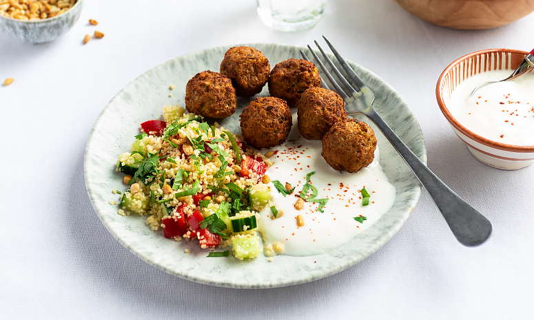 Falafel med couscous, grønnsaker og yoghurtdressing, servert på en tallerken med en gaffel liggende på høyre side.