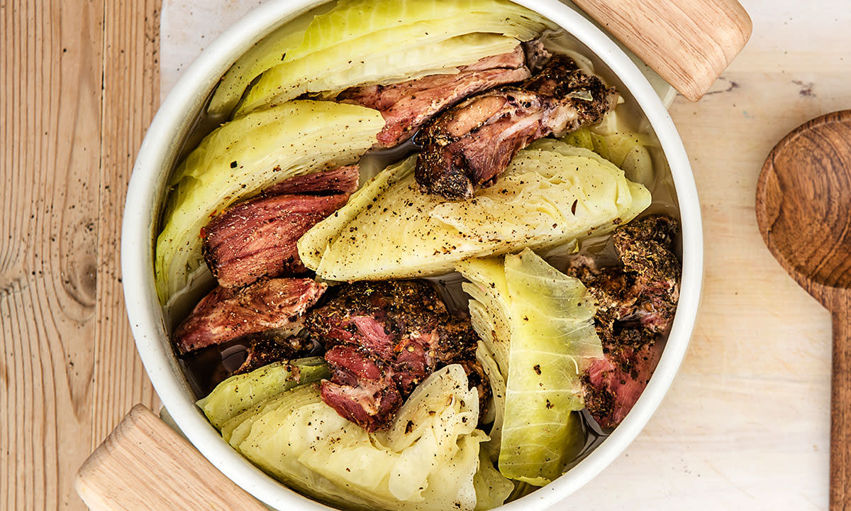 Kjapp fårikål med langtidskokt lammeskank og kål servert i hvit gryte på lyst trebord. Tresleiv ved siden av.