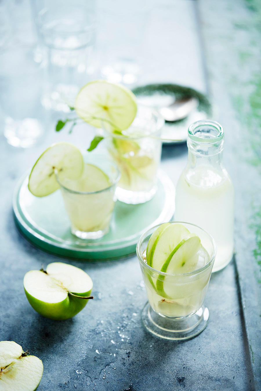 Eple- og ingefærlimonade servert i glass med grønne epler i skiver, på mørkt bord. Liten flaske med limonade, og halve grønne epler ved siden av.