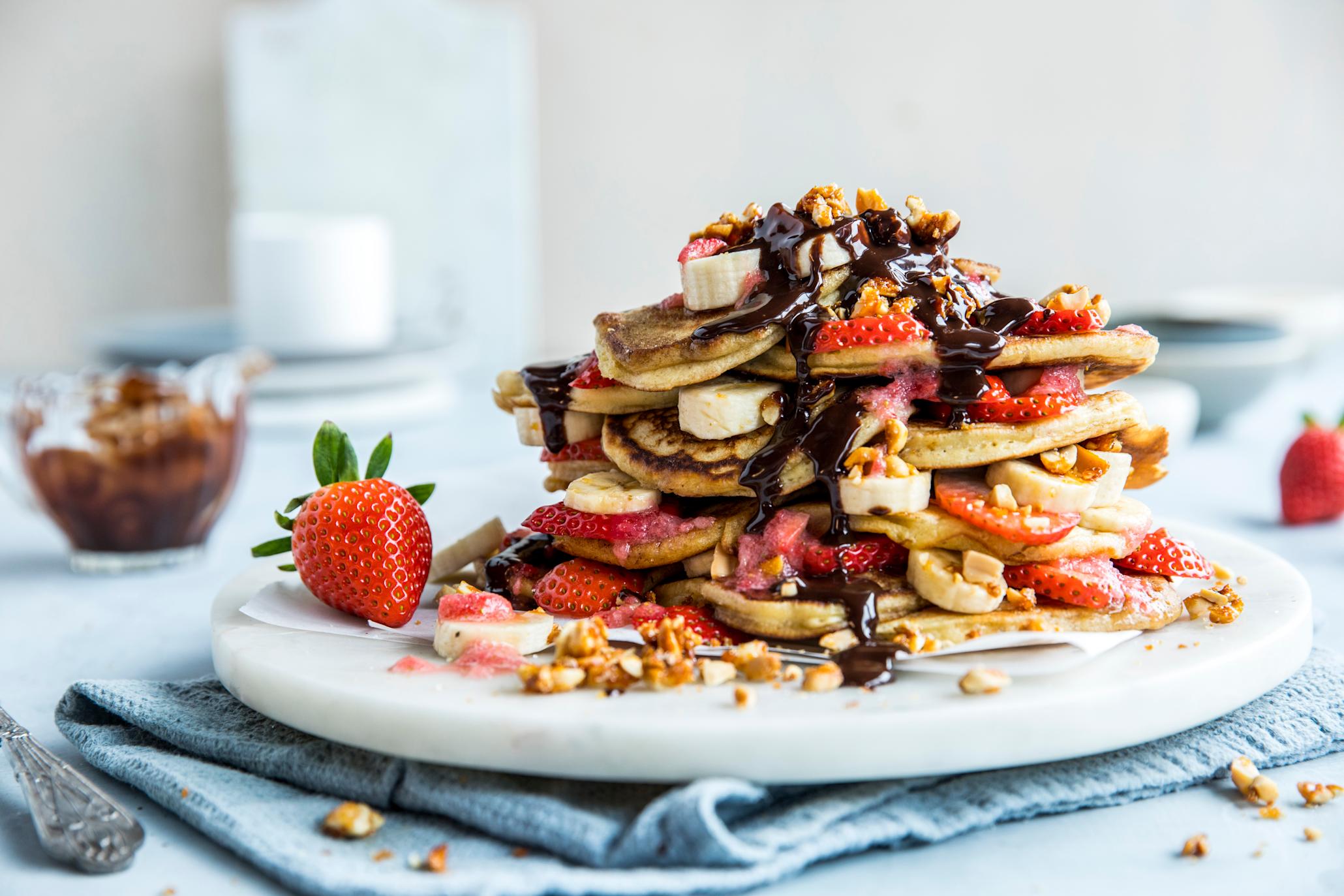 Glutenfrie bananpannekaker med hjemmelaget sjokoladesaus og karamelliserte peanøtter servert i tårn med jordbær- og bananskiver mellom, på hvitt rundt marmorfat. Liten skål med sjokoladesaus i bakgrunnen.