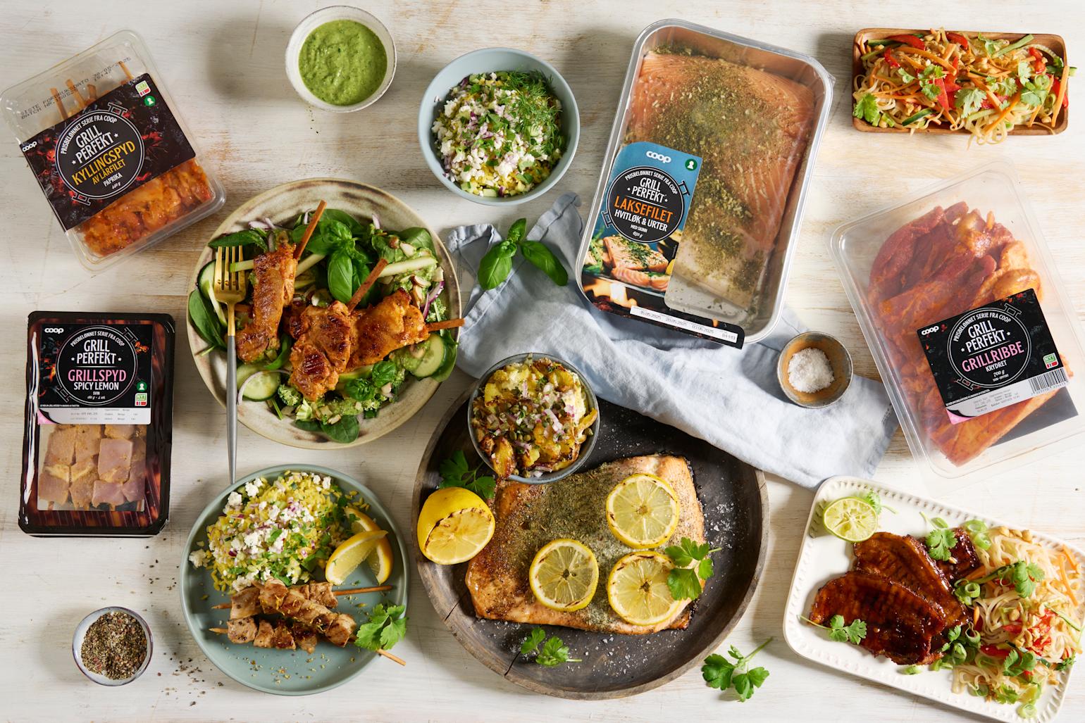Coop Grill Perfekt kjøtt, kylling og laks tilberedt og i forpakning, servert på forskjellig fat og tallerkner med tilbehør på lyst trebord.