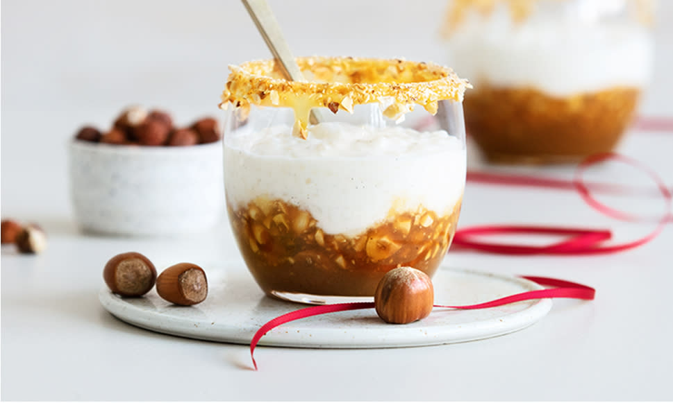 Riskrem servert i dessertglass med salt karamellsaus og røstede hasselnøtter. Glasset står på liten hvit tallerken på bord med små skåler hasselnøtter og karamellsaus i bakrunnen.