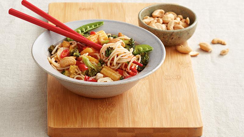 Sursøt vegetarwok med nudler, sukkererter, babymais, paprika og cashewnøtter i lys skål med røde spisepinner.