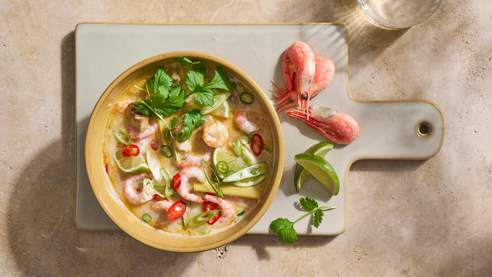Tom Kha Gai - thaisuppe med reker. Tom Kha Gai pyntet med frisk chili, vårløk og koriander, servert i lys skål på keramikkfjøl. Reker, limebåter og frisk koriander på siden.