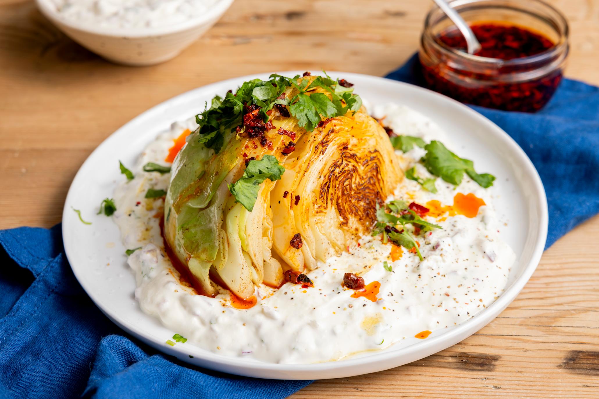Stekt hodekål med hjemmelaget yoghurtdressing servert på hvit tallerken med masse fersk koriander, og chili crisp på toppen. Tallerkenen står på et mørkeblått kjøkkenhåndkle på et lyst trebord, med skåler med chili crisp og yoghurtdressing ved siden av.