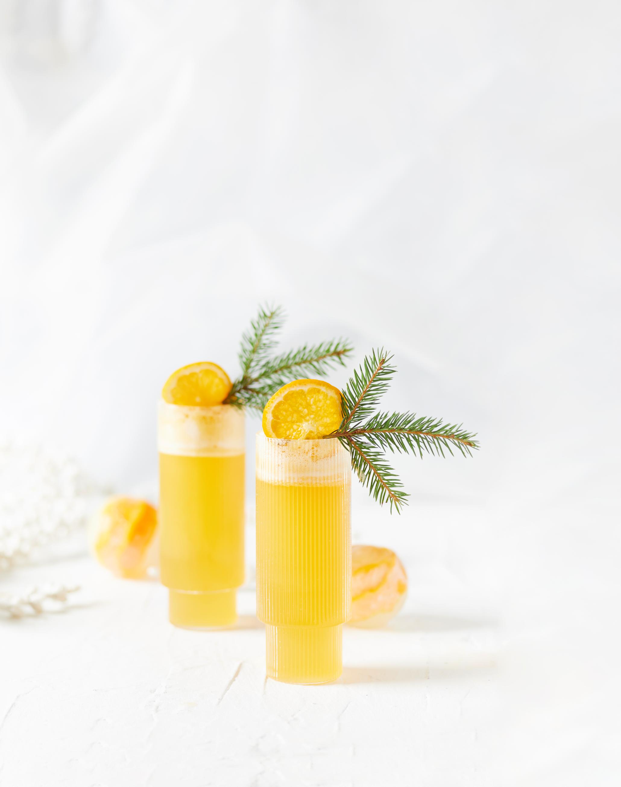 To høye glass med mocktailen Klementini, pyntet med klementinskiver og små kvister granbar. Glassene står på hvitt bord, med runde isbiter med klementinskall i bakgrunnen.