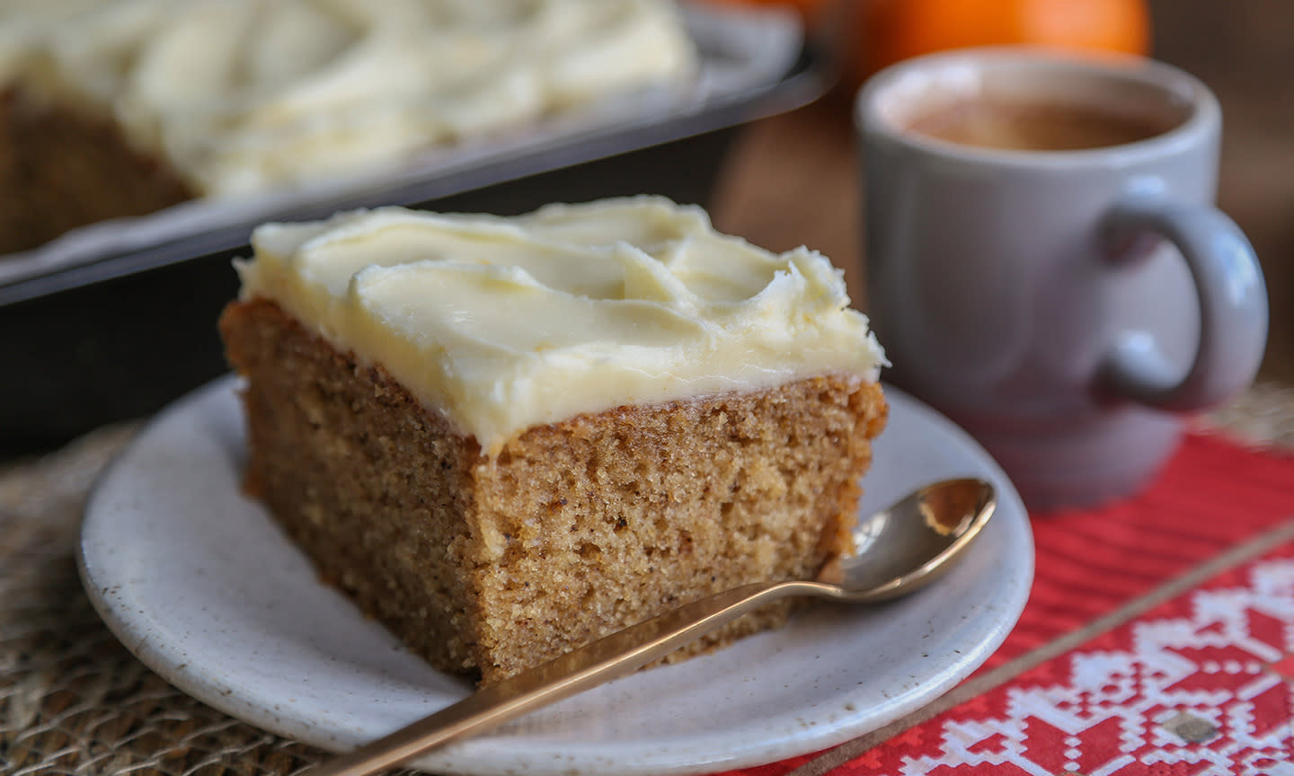 Et stykke krydderkake med kremostglasur servert på lys asjett på julepyntet bord, med en kopp kakao ved siden av og klementiner i bakgrunnen.
