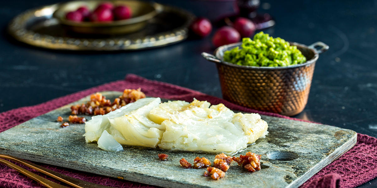 Et stykke ovnsdampet lutefisk servert på steinfjøl med sprøstekt bacon. Liten messingskål i bakgrunnen med hjemmelaget ertepuré.