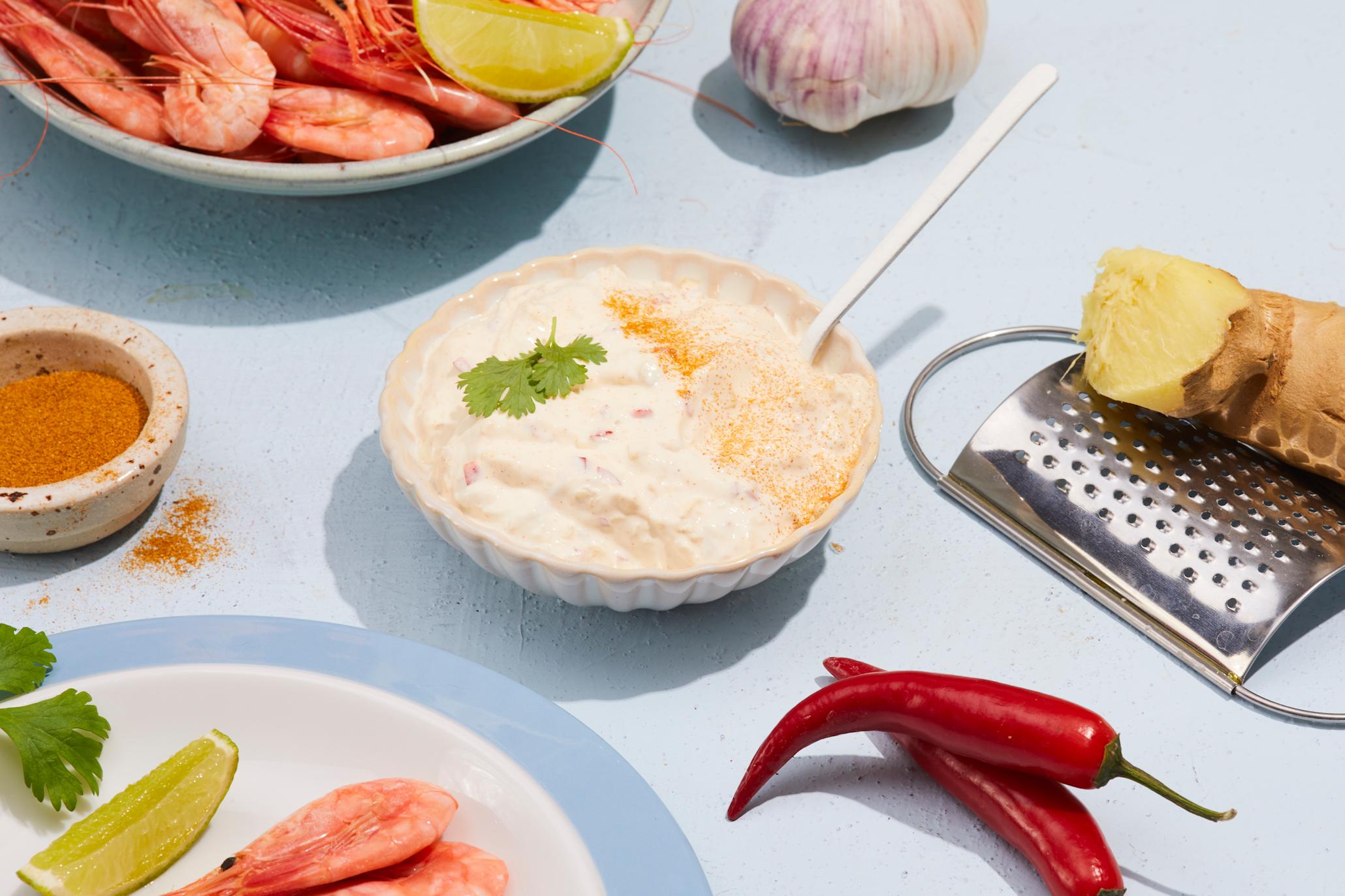 Kremet dipsaus med asiatiske smaker servert i liten lys skål på blått bord med ferske reker, chili, hvitløk, ingefær, limebåter og friske urter.