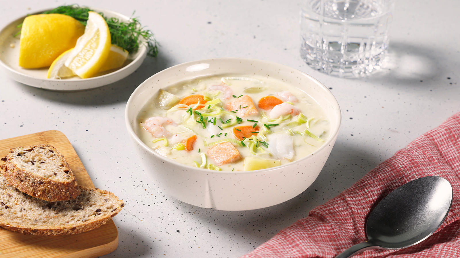 Bergensk fiskesuppe med laks og grønnsaker servert i lys skål på dekket middagsbord med brødskiver, sitron og dill på siden.