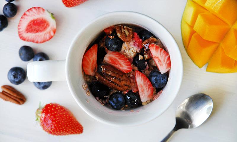 Rask havregrøt laget i mikroen, servert i hvit kopp, med ferske jordbær, blåbær, pekannøtter og honning ringlet over. Hvit bakgrunn, med ferske bær og mango på siden.