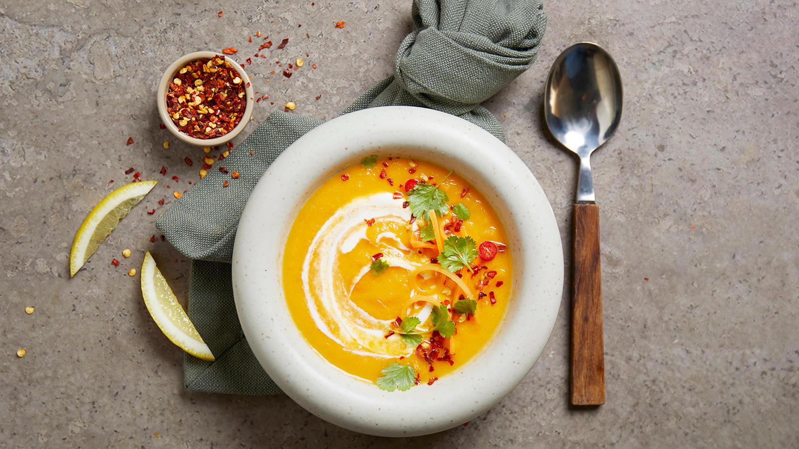 Kremet gulrot- og ingefeæsuppe servert i hvit skål med strimlet gulrot, chili og koriander på toppen. Sitronbåter og liten skål med chiliflak på siden.