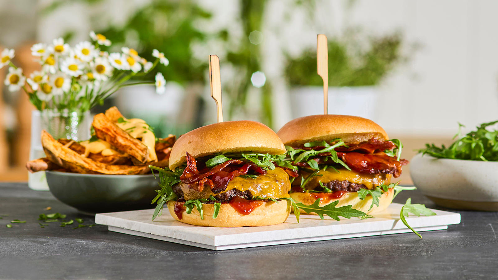 Maple baconburger med loaded søtpotetfries servert på hvitt steinfat med en bolle søtpotetfries i bakgrunnen.