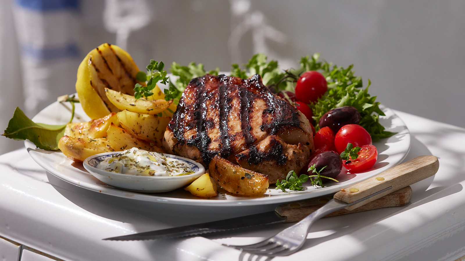 Greske svinekoteletter og ovnsbakte poteter med sitron servert på hvit asjett med grønn salat, tomater og tzatziki.