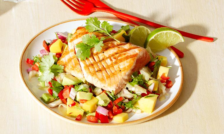 Ett stykke grillet laks med sitrusmarinert avokado- og mangosalat, servert på hvit tallerken med rødt bestikk ved siden av.