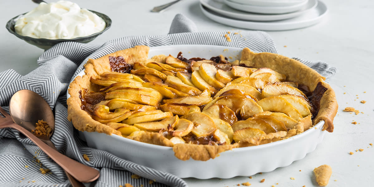 Eplepai med karamellsaus servert i hvit paiform på hvitt bord, med stripete kjøkkenhåndkle og messingfarget bestikk ved siden av. Mørk skål med pisket krem i bakgrunnen.