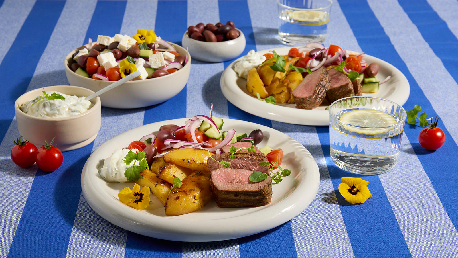 Urtemarinert fårefilet med greske sitronpoteter servert på lyse talerkner på blåstripet duk. Skåler med hjemmelaget tzatziki og gresk salat i bakgrunnen.