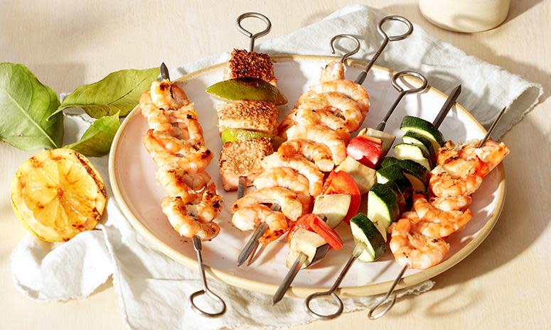 Grillspyd med laks og marinert scampi, ved siden av grillspyd med squash og paprika, på hvit tallerken. Lys bakgrunn, grillet sitron ved siden av.