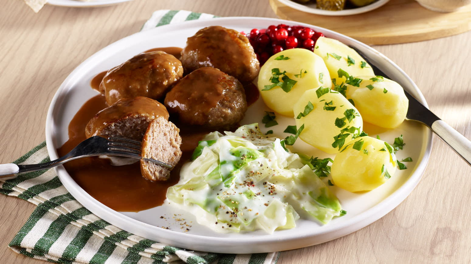 Kjøttkaker med kålstuing, poteter og tyttebærsyltetøy servert på hvit tallerken på dekket middagsbord.