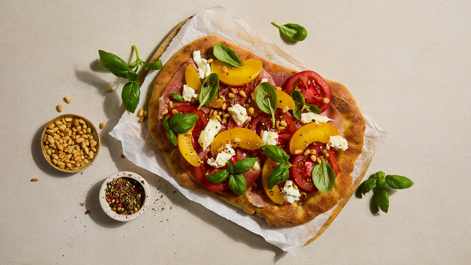 Pinsa pizza med spekeskinke, tomat og fersken pyntet med frisk basilikum og pinjekjerner, og servert på lys fjøl med bakepapir.