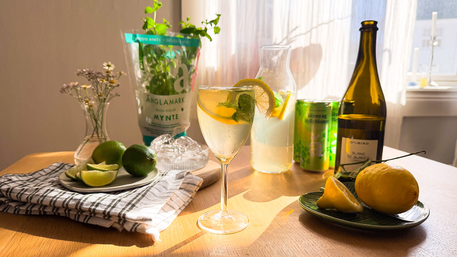 Hugo Spritz Mocktail servert i vinglass med sitronskiver og mynte. Vinglasset står på et kjøkkenbord med sitrusfrukter, ferske urter og Änglamark drikkebokser.